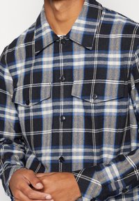 Koszula w kratę w kolorach granatowym, niebieskim i beżowym, wykonana z teksturowanej tkaniny. Posiada dwie kieszenie na piersi oraz czarne guziki, zaprojektowana z kołnierzykiem.