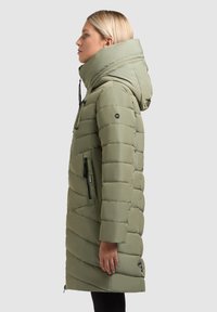 Gevoerde lange jas in licht olijfgroen met een grote capuchon, ritszijzakken en een gewatteerde textuur met een chevronpatroon.
