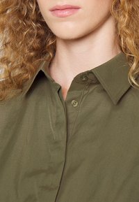 Camicia verde oliva con colletto strutturato, texture liscia e chiusura a singolo bottone sulla parte superiore della patta.