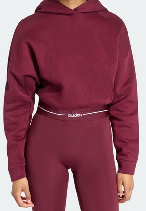 Kvinde iført vinrød Adidas hættetrøje og højtaljede leggings med hvidt Adidas-logo på taljen, står med afslappede hænder.