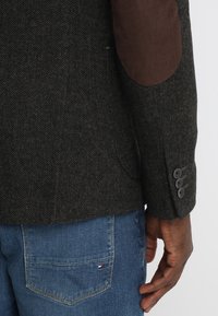 Sak s tmavým herringbone vzorem, hnědými semišovými loketními záplaty, čtyřmi knoflíky na manžetách, kombinovaný s modrými džíny a logem na zadní kapse.