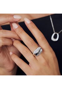 Silver ring med texturerad band och slät centraldel, bärs på en hand med manikyrerade naglar, vilket framhäver dess glänsande yta och design.