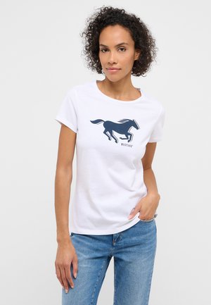 Woman with curly hair wearing white t-shirt with black running horse graphic and blue jeans, posing with one hand in pocket.