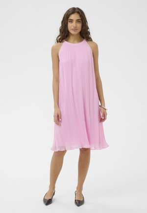 Žena kučeravých vlasov nosí ružové plisované šaty bez rukávov po kolená a čierne špicaté lodičky, stojí pred jednofarebným pozadím.