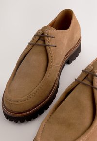 Brune ruskindsko med rundet tå, snøredesign, kontrastsyning og robuste gummisåler. Nærbillede, der viser tekstur og form.