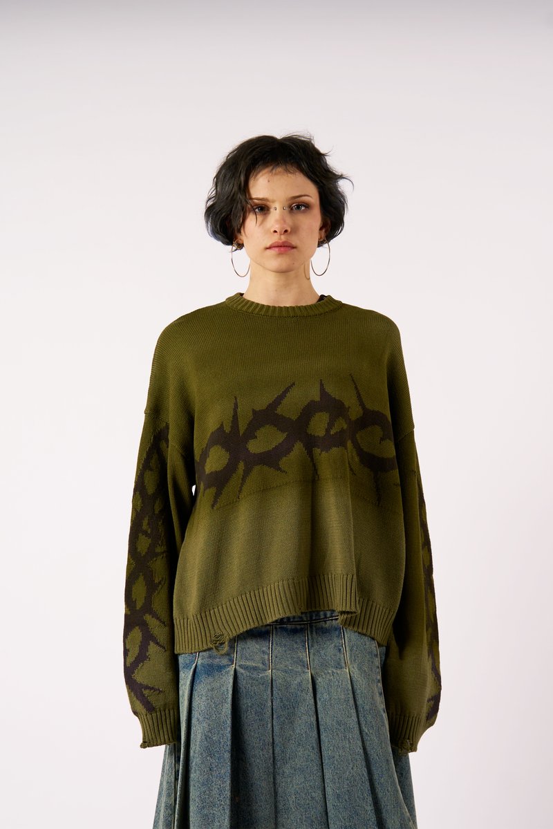 Ελιά πράσινος πλεκτός σκούφος με σκούρο καφέ αφηρημένο σχέδιο, ραβδωτός λαιμός και μακριά μανίκια, συνδυασμένος με πλισέ τζιν φούστα.