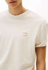 T-shirt in cotone beige con colletto rotondo, maniche corte arrotolate e una piccola etichetta con logo nella parte superiore sinistra che presenta "TOMMY JEANS" in caratteri testurizzati.