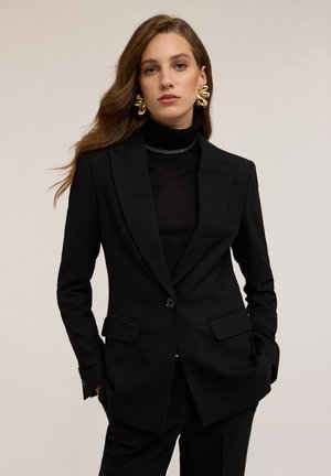 Vrouw gekleed in een oversized zwarte blazer en broek, zwarte coltrui, grote gouden statement oorbellen, staande met handen in de zakken.