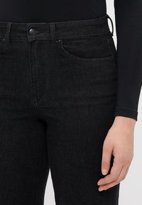 Fekete farmernadrág, szűkített szabással, elülső gombos záródással, standard zsebekkel és sima textúrával.