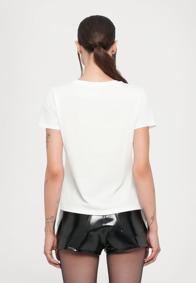 Weißes Baumwoll-T-Shirt mit kurzen Ärmeln, das einen Rundhalsausschnitt und einen lockeren Schnitt aufweist. Das Modell kombiniert es mit glänzenden schwarzen Shorts.