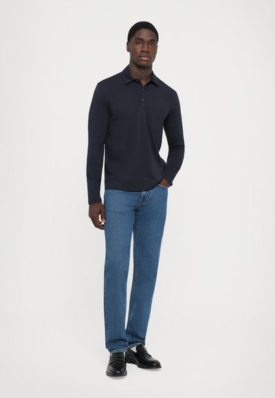 Man standing against white background wearing a dark long-sleeve polo shirt, blue jeans, and black dress shoes, with one hand in pocket.