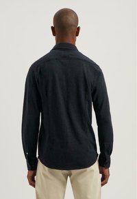 Hombre con cabello corto, vistiendo una camisa de manga larga de color azul marino oscuro y pantalones beiges, de pie de espaldas contra un fondo claro y liso.