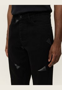 Pantalones de mezclilla negra con un corte regular, costuras visibles y parches triangulares con acentos de logo en la parte delantera y el muslo.