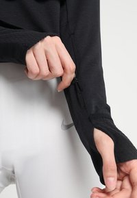 Haut noir à manches longues avec des trous pour les pouces, montrant des poignets texturés. Leggings blancs ajustés avec un logo Nike argenté sur la hanche.