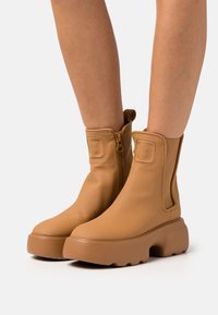 Hellbraune Knöchelstiefel aus glattem Leder mit seitlichem Reißverschluss, elastischen Einsätzen und einer klobigen Plateausohle. Schlichtes Design ohne sichtbare Muster.