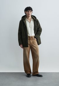 Kaki parka z ruto, bež pulover z zadrgo in široke rjave hlače. Črni čevlji dopolnjujejo opravo. Gladke teksture in sproščen kroj.