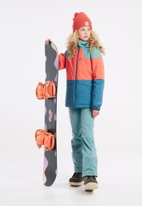 Plava i narančasta snowboard jakna preko tirkiznih hlača, s crnim snowboardom koji ima narančaste vezove i apstraktne uzorke. Narančasta kapa.