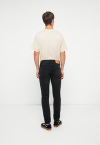 Heren lichte beige t-shirt, zwarte slim-fit jeans en zwarte sneakers met witte details; zichtbaar leren label op de achterkant van de tailleband van de jeans.
