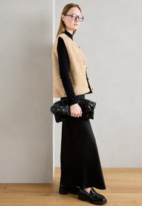 Beige konstpälsväst över en svart rullkrage, parat med en svart ribbad clutch och en lång svart kjol. Glansiga svarta loafers fulländar looken.
