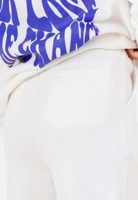 Vita mjukisbyxor med en slät textur och en bakficka. Del av en vit outfit som innehåller en grafisk sweatshirt med blått tryck.