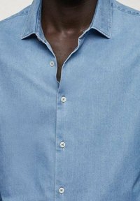 Jasnoniebieska koszula dżinsowa z kołnierzykiem, zasuwaną na guziki i białymi guzikami. Tkanina ma gładką teksturę i subtelny połysk.