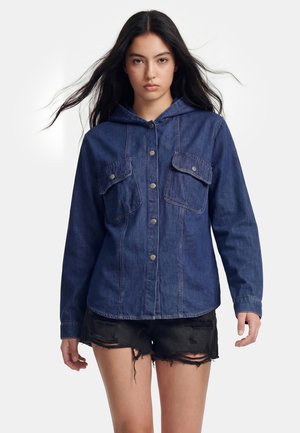 Woman with long dark hair wearing a buttoned dark blue denim jacket with flap pockets and black ripped shorts against a plain background.