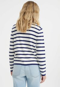 Pullover a righe bianco e blu navy, con maniche lunghe, scollatura rotonda e polsini a costine. Jeans azzurri e vista posteriore visibile.