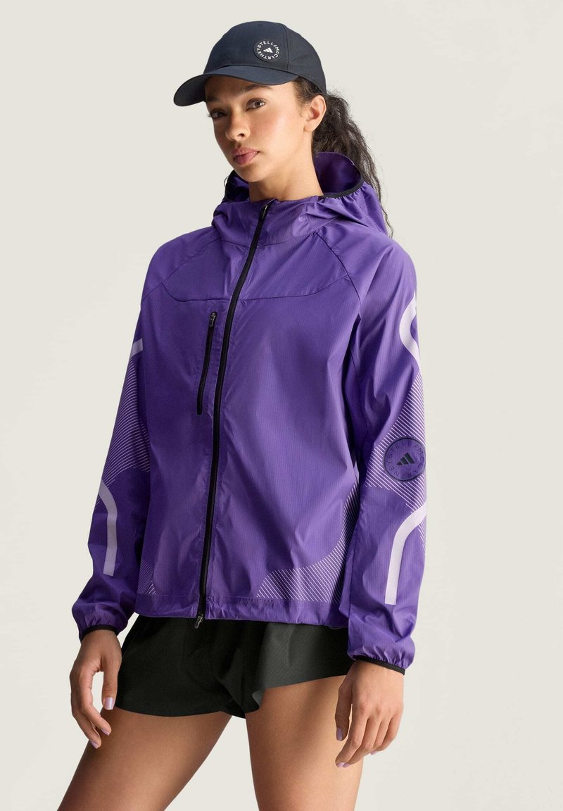 Chaqueta ligera púrpura con capucha ajustada, cremallera frontal y detalles reflectantes. Presenta un parche de logo en el brazo izquierdo y patrones a rayas.