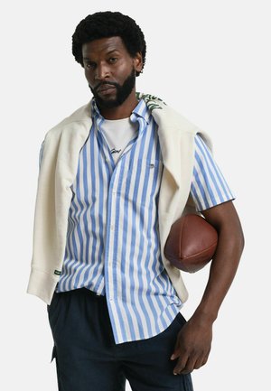 Short-sleeve button-up shirt with blue and white stripes, layered under a cream crewneck sweater, holding a brown football.