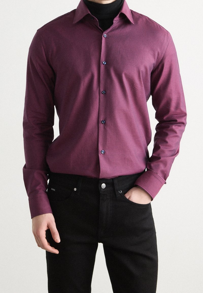 Homme portant une chemise violette boutonnée sur un col roulé noir avec un pantalon noir, une main dans la poche, sur fond uni.