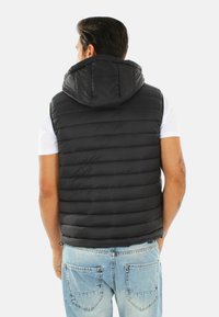 Gilet piumino nero con cappuccio, caratterizzato da cuciture orizzontali e una superficie liscia. Indossato sopra una t-shirt bianca, abbinato a jeans in denim chiaro.