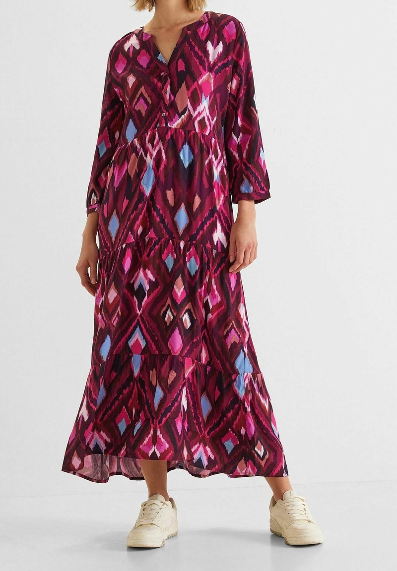 Femme portant une longue robe à motifs géométriques multicolores, manches trois-quarts et baskets blanches sur un fond uni.