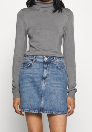 Femme portant un pull à col roulé gris à manches longues et une jupe en denim bleue longueur genoux, debout devant un fond uni.