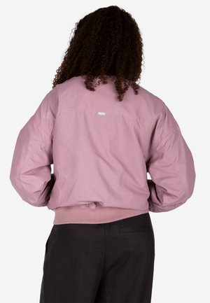 Person mit lockigem Haar, die eine lockere rosa Jacke und schwarze Hose trägt und mit dem Rücken zu einem einfarbigen Hintergrund steht.