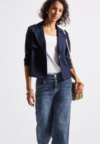 Marinblå kort jacka med slag och uppvikta ärmar, tillsammans med ljusblå vida jeans. Modellen håller en liten beige axelremsväska.