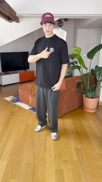 Mustapaita, jossa on lyhyet hihat ja pyöreä logo rinnassa, yhdistettynä harmaisiin leveälahkeisiin housuihin ja hopeisiin lenkkareihin nykyaikaisessa sisätilassa.