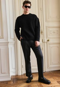 Zwarte fuzzy trui gecombineerd met gestructureerde grijze broek en zwarte plateauschoenen. Model staat met de hand in de zak tegen een pastelmuur.