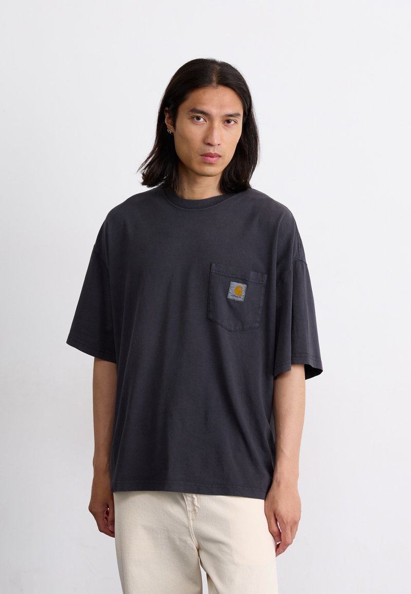 T-shirt surdimensionné gris foncé en coton. Col rond, manches courtes, poche frontale avec un patch logo. Porté avec un pantalon de couleur claire.