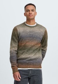 Suéter de punto con cuello redondo que presenta un patrón en degradé de marrones y verdes con un acabado texturizado, combinado con una camiseta blanca y vaqueros oscuros.