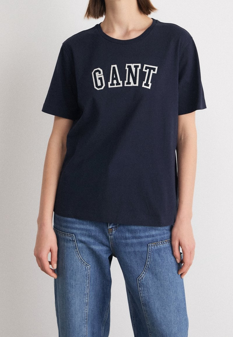 Navyblauwe katoenen T-shirt met korte mouwen, ronde halslijn en wit "GANT" logo over de borst. Gecombineerd met lichtblauwe jeans.