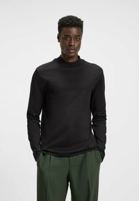Schwarzes Langarmshirt mit hohem Kragen, aus glattem Stoff. Kombiniert mit grünen maßgeschneiderten Hosen in legerem Schnitt.