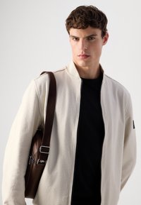 Giovane uomo che indossa una giacca beige con zip sopra una maglietta nera, mentre porta una borsa a tracolla in pelle marrone su uno sfondo semplice.