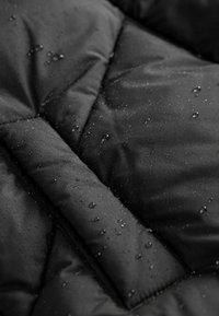 Tissu imperméable noir avec des gouttes d'eau perlées sur sa surface, montrant une couture matelassée et une texture douce et rembourrée.