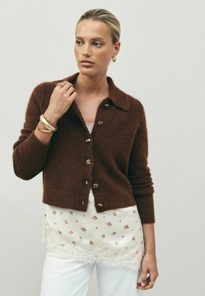 Frau trägt eine braune, geknöpfte Strickjacke über einem weißen Top mit floraler Spitzenverzierung und weißen Hosen, mit goldenen Creolen und Armbändern.