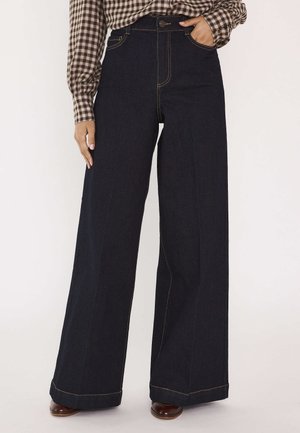 Jeans blu scuro a vita alta con gamba larga e cuciture gialle visibili, indossati con scarpe marroni e una camicia a quadri marrone a maniche lunghe.