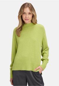 Pull vert avec un col montant, texture tricotée, poignets et ourlet côtelés, et coupe ample. Porté avec un pantalon en tissu sombre et lisse.