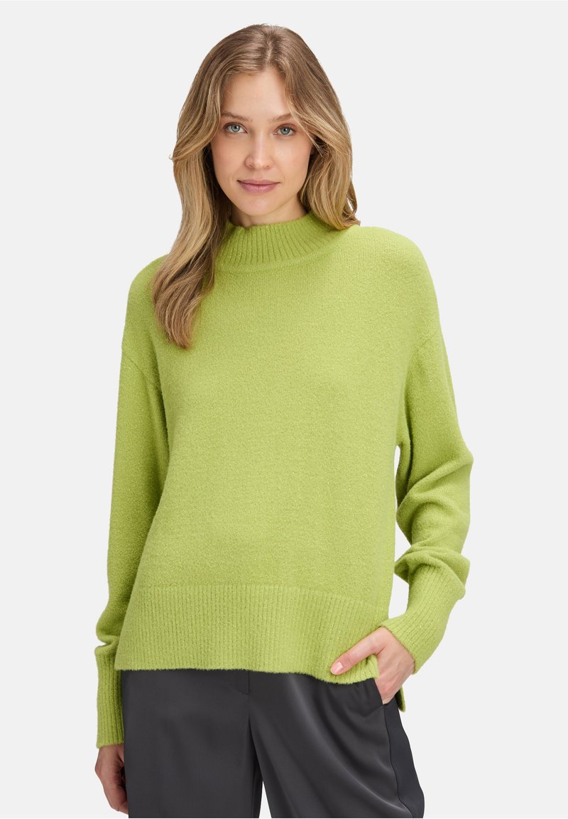 Pull vert avec un col montant, texture tricotée, poignets et ourlet côtelés, et coupe ample. Porté avec un pantalon en tissu sombre et lisse.