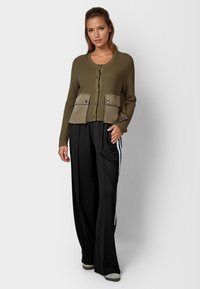 Olivgrüner gerippter Cardigan mit Knopfverschluss und zwei vorderen Taschen; getragen mit schwarzen weit geschnittenen Hosen, die weiße Seitenstreifen haben.