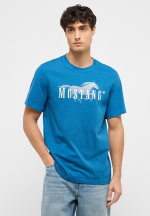 Mann trägt blaues Mustang-T-Shirt und hellblaue Jeans, steht mit einer Hand in der Tasche vor weißem Hintergrund.