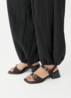 Πόδια που φορούν καφέ σανδάλια με τακούνι τύπου block από σουέντ και ασημένιες αγκράφες, συνδυασμένα με άνετα μαύρα παντελόνια με μανσέτες σε λευκό φόντο.